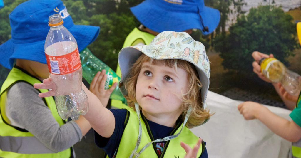 Child dressed in floopy hat and yellow high viz top hots up a plastic bottle.